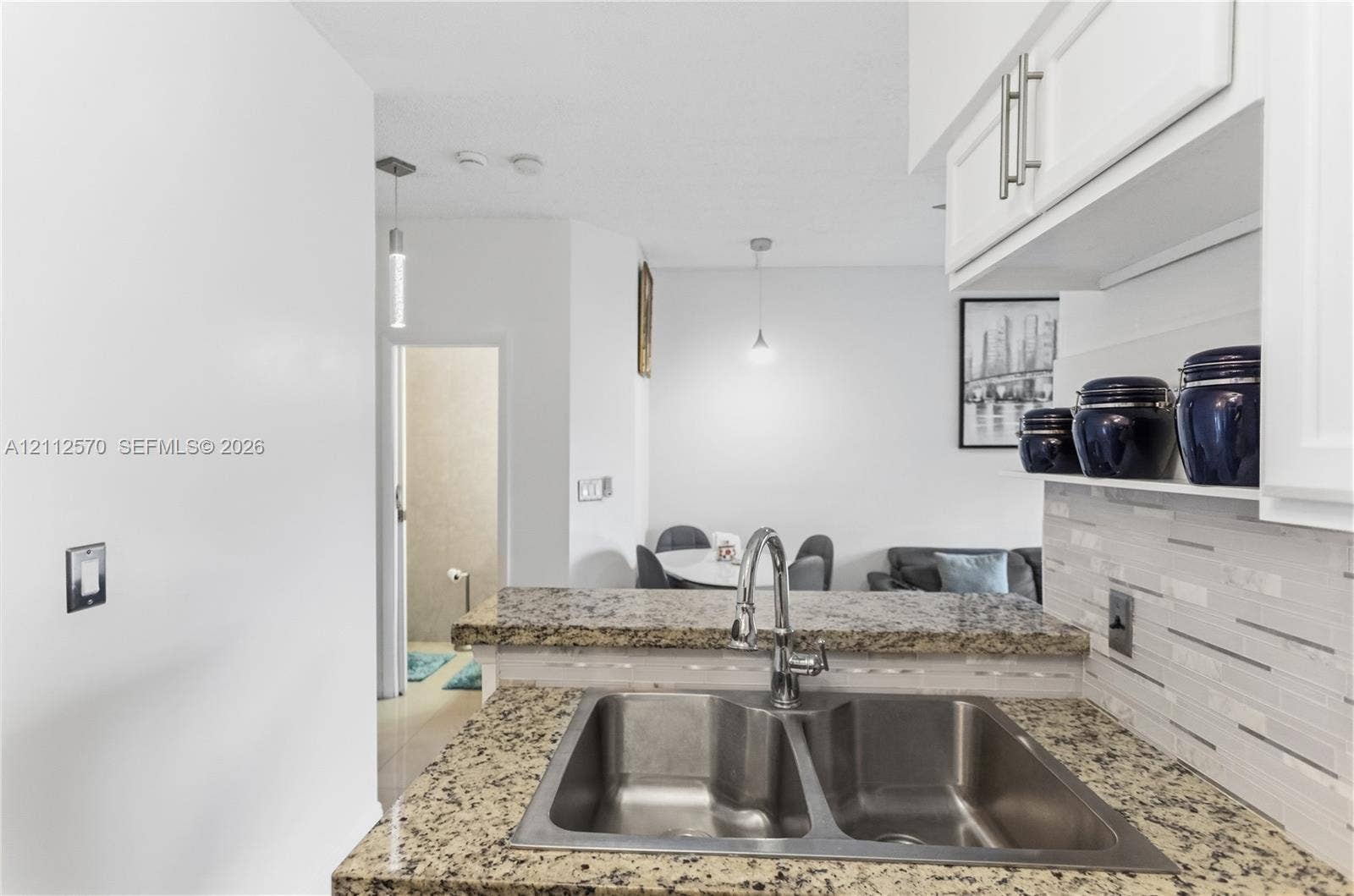 Kitchen featuring stone countertops, a dual-basin stainless steel sink, polished chrome faucet, white cabinetry, and a multi-toned linear tile backsplash