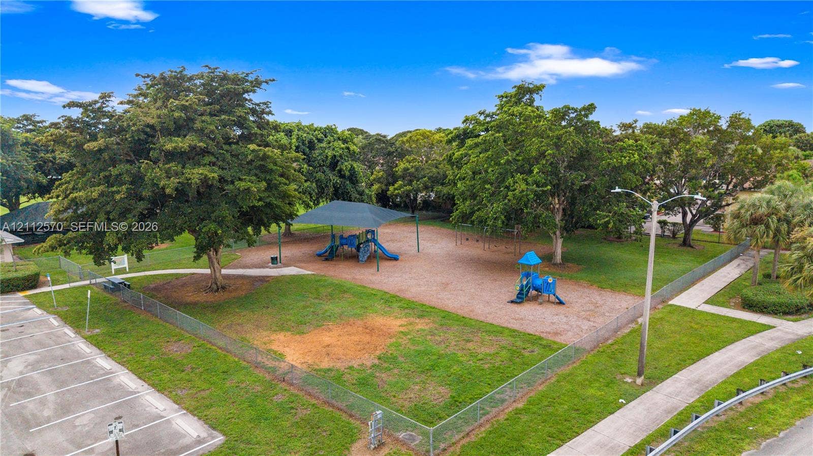 Outdoor recreational area featuring a covered playground structure, open green space, mature trees, and perimeter fencing