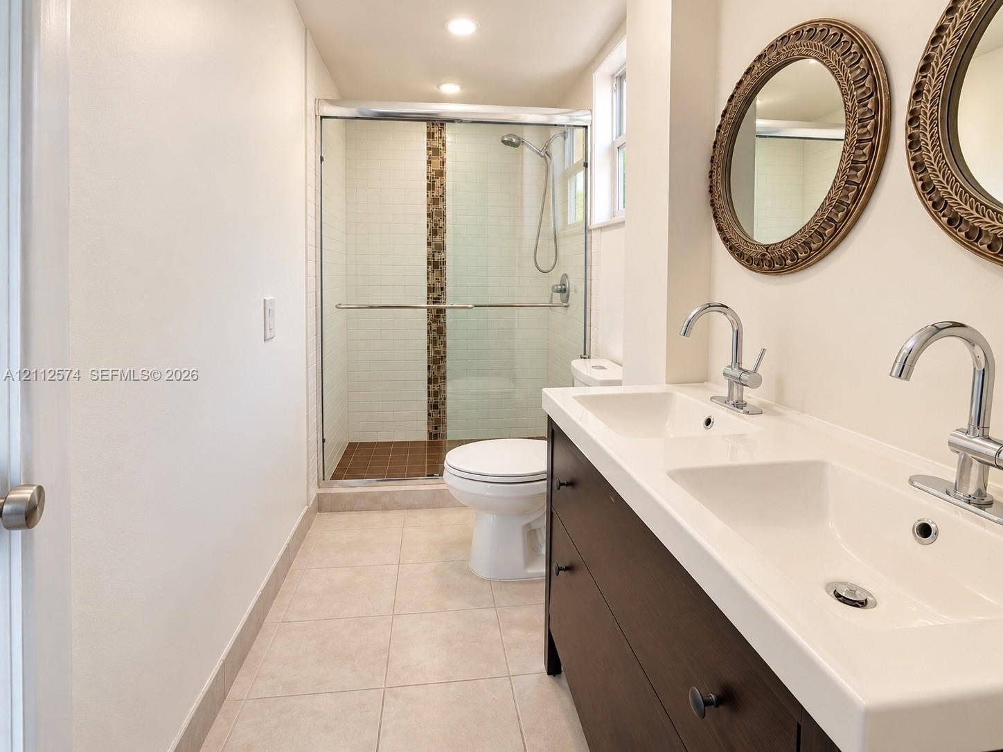 Dual-sink vanity with a dark wood-finish cabinet and integrated countertop, glass-enclosed walk-in shower with tile surround, and neutral floor tiling