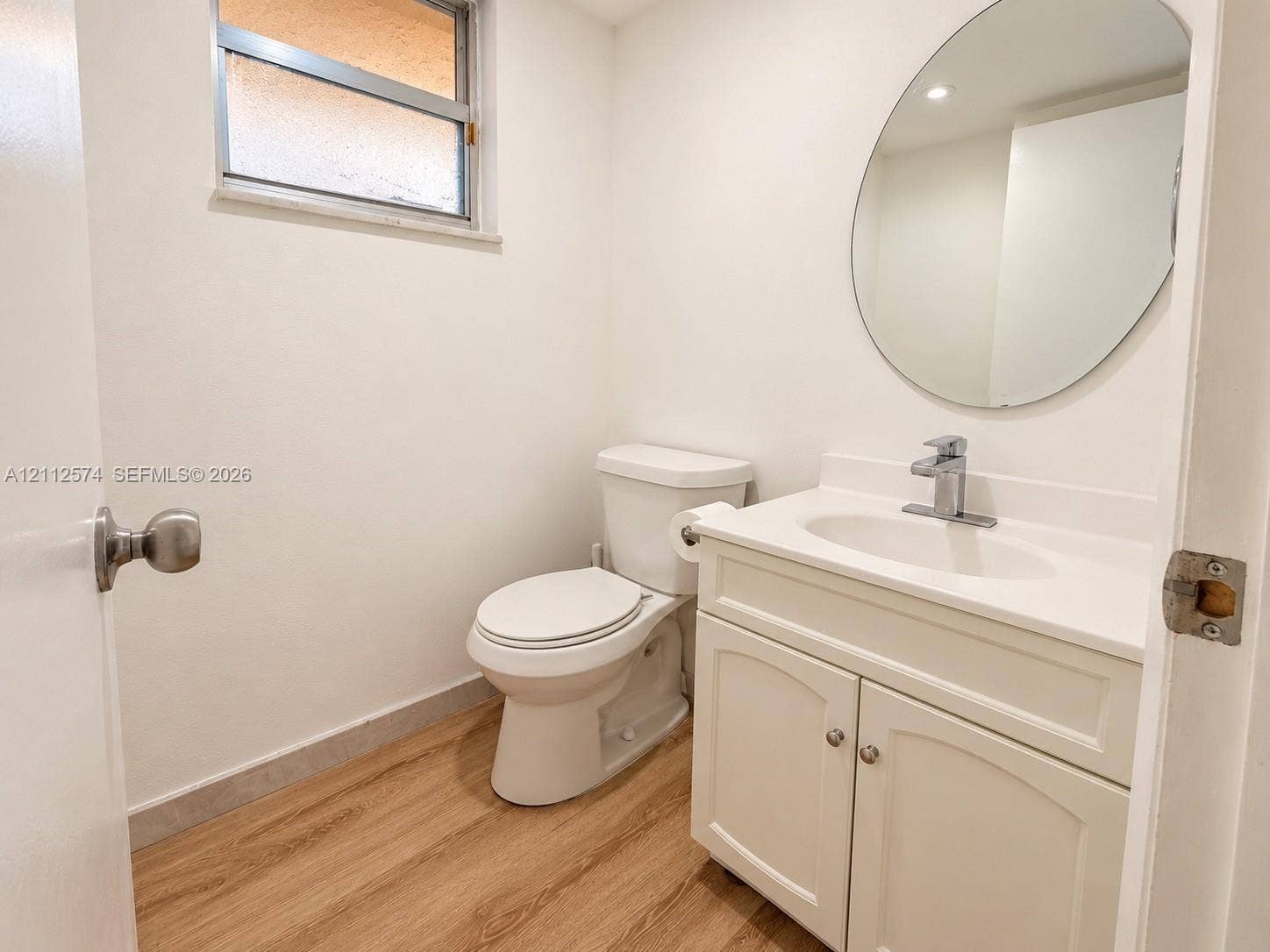 White vanity with a molded countertop and basin, chrome faucet, oval wall-mounted mirror, two-piece toilet, and wood-finish flooring