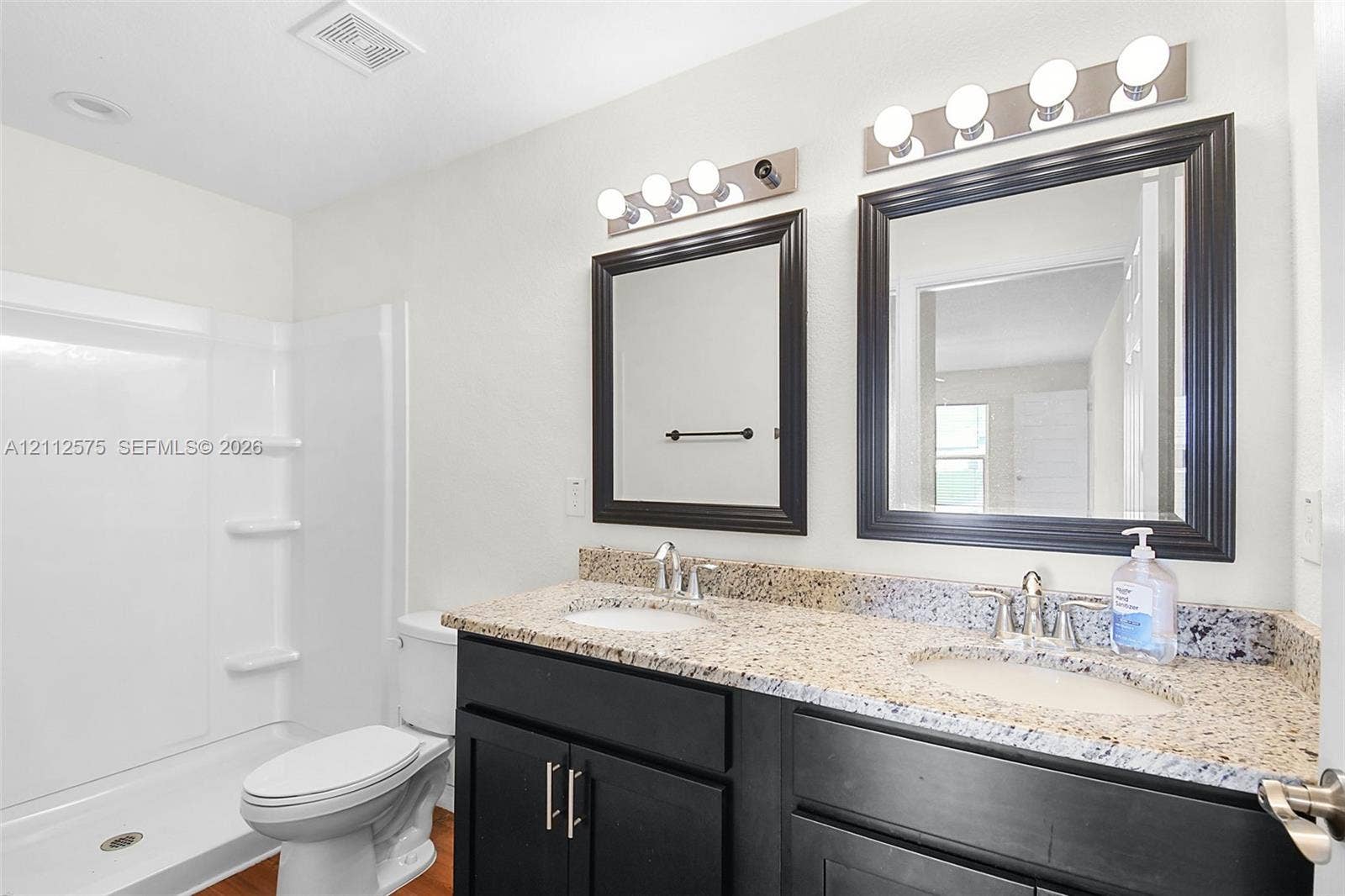 9041 E Broad, Labelle, FL 33935 Dual-sink vanity with a stone-finish countertop, dark cabinetry, matching wall-mounted mirrors, and dual four-light fixtures, accompanied by a fiberglass shower surround