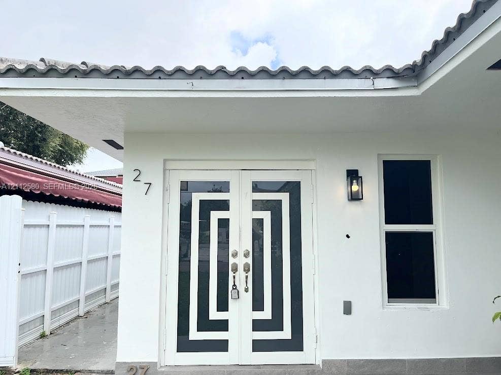 White stucco exterior featuring a double-door entry with geometric detailing, tile roof, vertical single-hung window, and wall-mounted exterior lighting