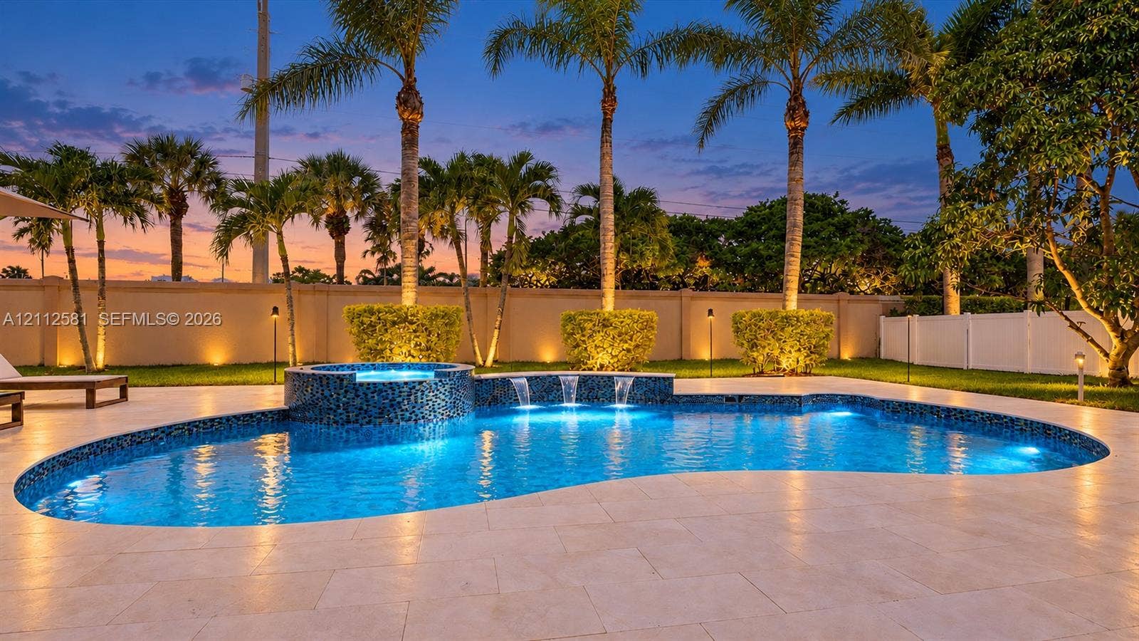 Custom-designed swimming pool featuring a tiled spa, waterfall accents, and large-format stone pavers, set within a landscaped perimeter featuring mature palm trees and a privacy wall