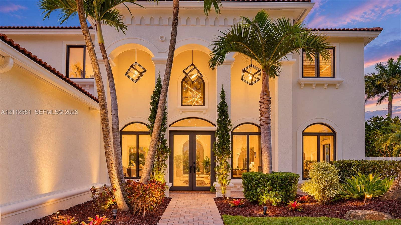 Stucco exterior featuring an arched entryway with decorative corbels, dual-glass entry doors, multiple geometric hanging pendant lanterns, and arched window detailing