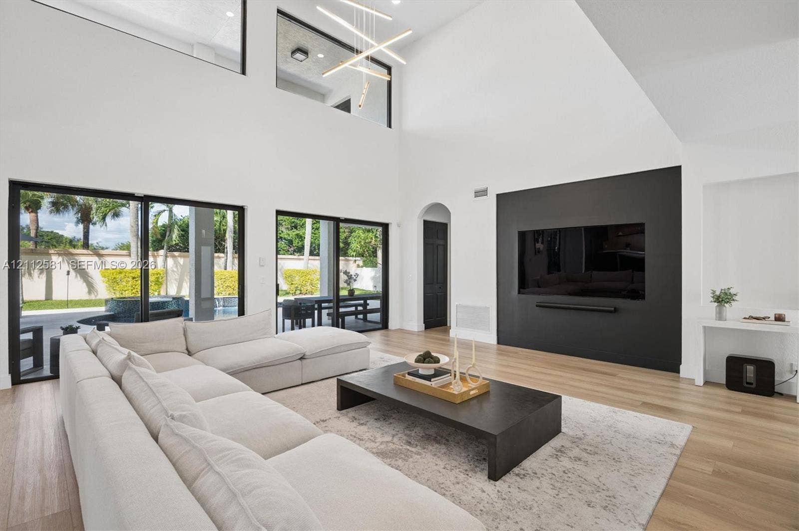Double-height living area featuring vaulted ceilings, floor-to-ceiling glass sliding doors, wood-finish flooring, a matte black accent wall with a built-in media recess, and a geometric chandelier
