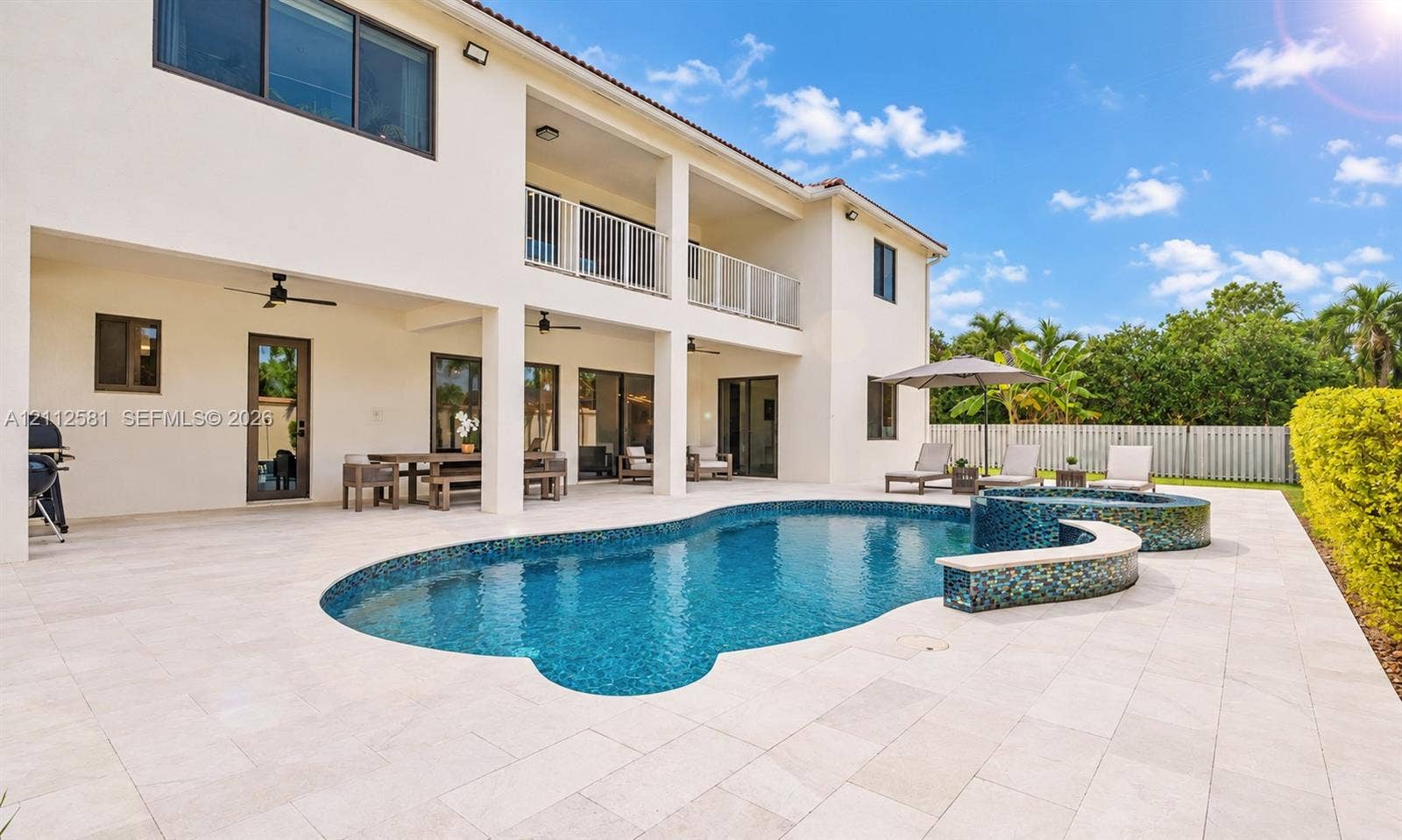 Rear exterior featuring a two-level covered patio, a free-form swimming pool with a mosaic tile spillover spa, and expansive stone-paver decking