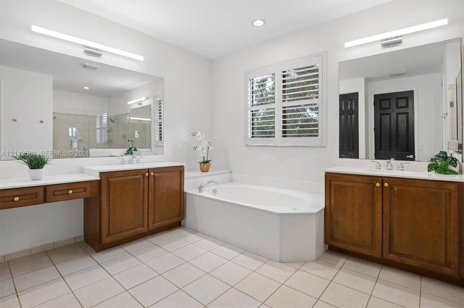 Dual wood-finish vanities with solid-surface countertops, soaking tub with tile surround, expansive wall-mounted mirrors, plantation shutters, and ceramic tile flooring