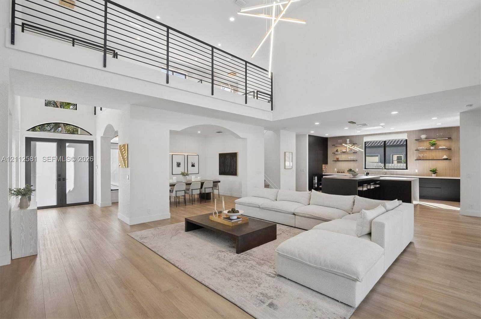Double-height ceiling with geometric pendant lighting, wood-finish flooring, metal horizontal-bar mezzanine railing, and kitchen featuring dark-tone cabinetry with open wood shelving