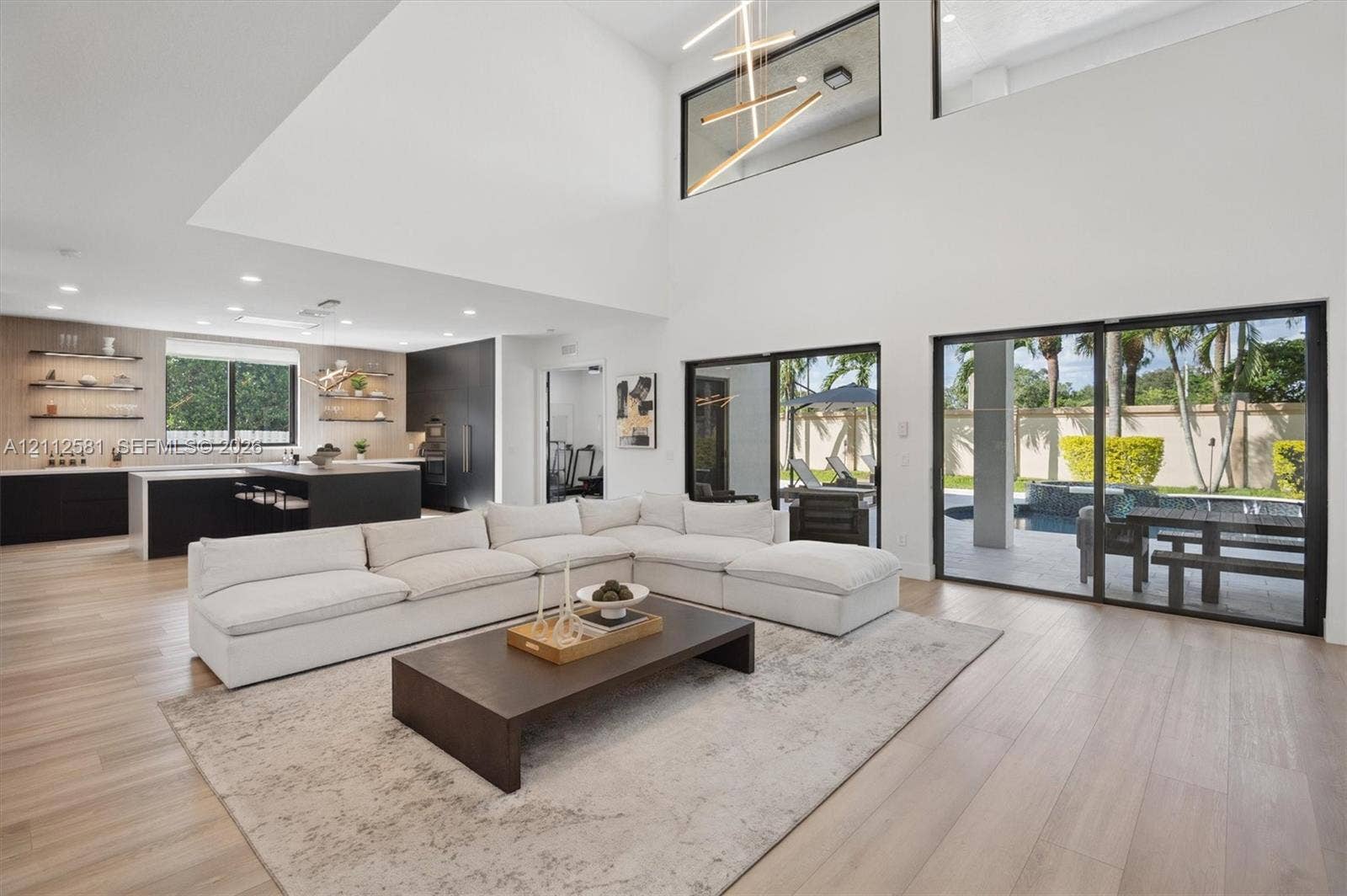 Double-height ceiling, expansive sliding glass doors, wood-finish flooring, recessed lighting, and kitchen featuring built-in cabinetry and floating shelving