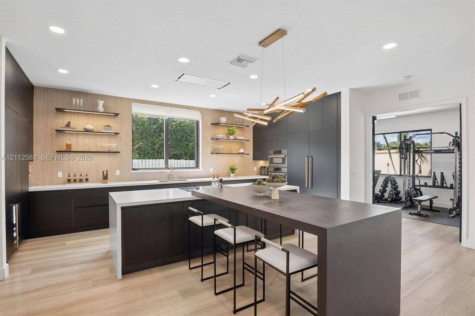 Contemporary kitchen featuring matte dark cabinetry, wood-finish wall paneling, integrated stainless steel appliances, quartz countertops, and a waterfall-edge island with built-in seating