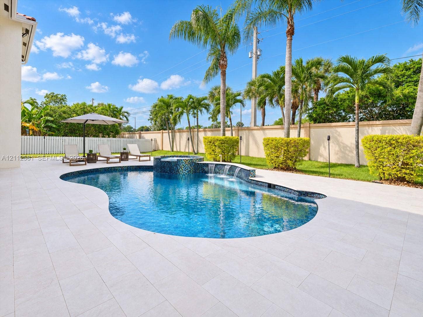 Free-form swimming pool featuring a tiled spa, waterfall feature, light-toned stone decking, and perimeter privacy wall