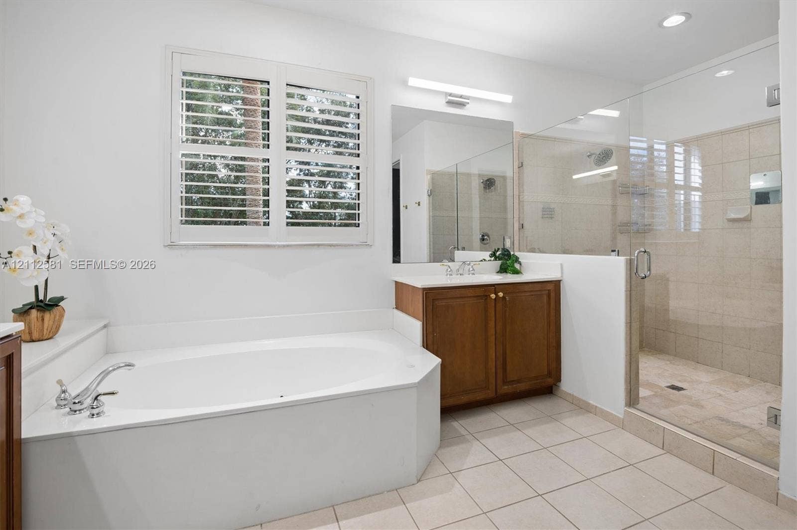 Built-in soaking tub with a deck-mounted faucet, wood cabinetry vanity, large wall mirror, glass-enclosed shower with tile surround, and plantation shutters