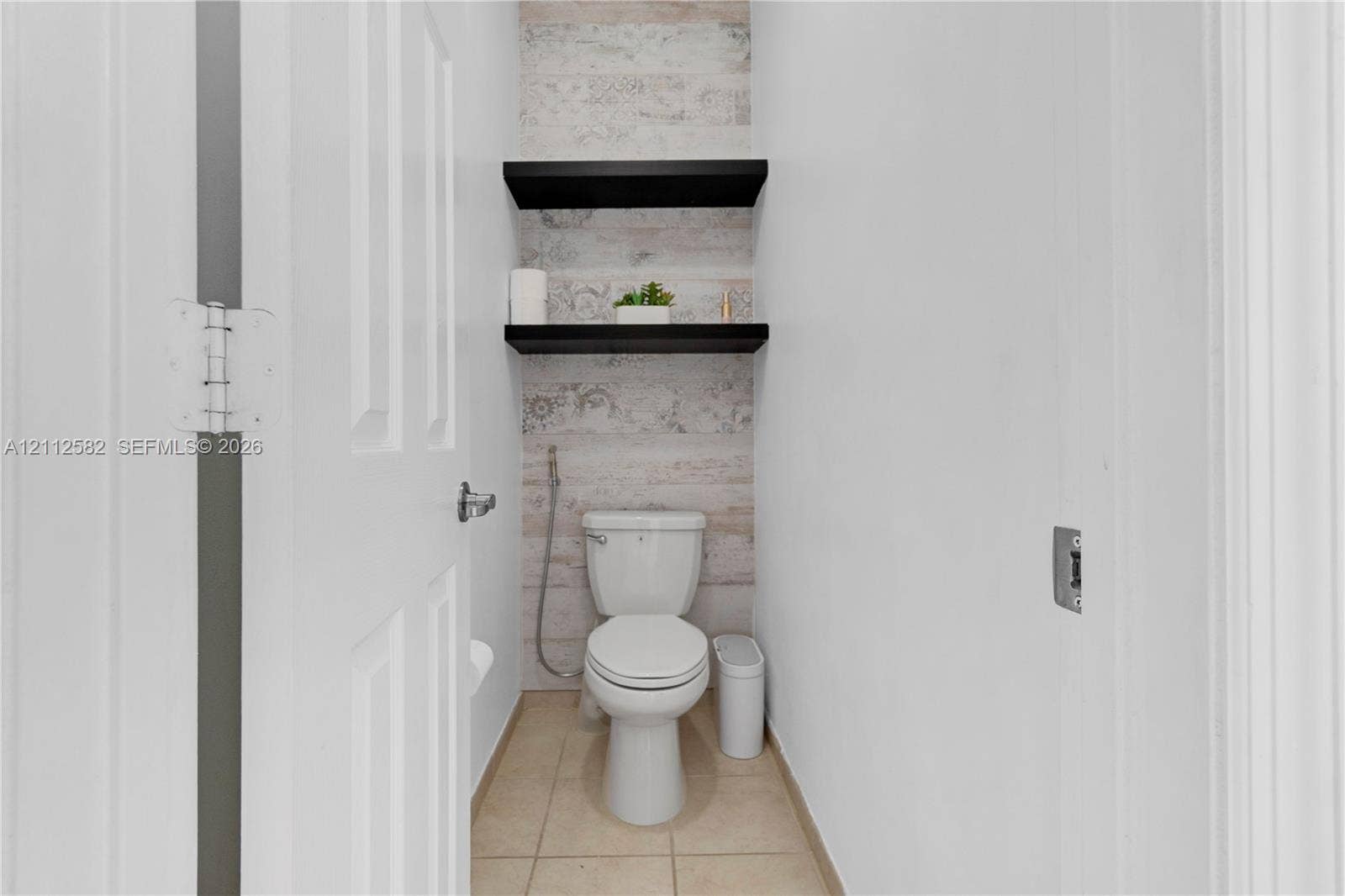 Restroom featuring ceramic tile flooring, two black floating shelves, decorative wall surfacing, and a wall-mounted bidet sprayer