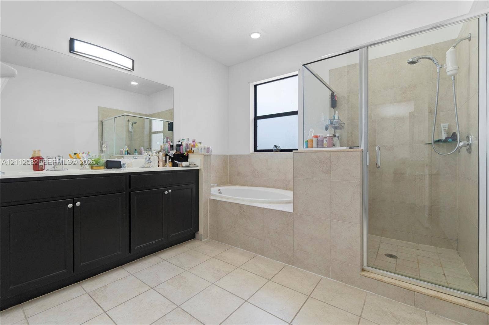 Dark cabinetry with dual undermount sinks, an expansive wall-to-wall mirror, a tiled deck-mounted soaking tub, and a separate glass-enclosed shower with tiled surround
