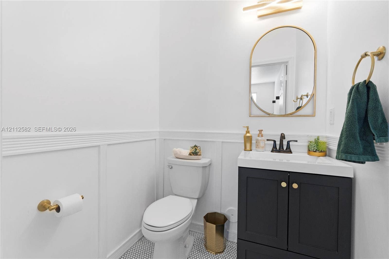 Powder room featuring wainscoting, a dark vanity cabinet with a stone-top sink, arched gold-rimmed mirror, patterned floor tile, and gold hardware fixtures