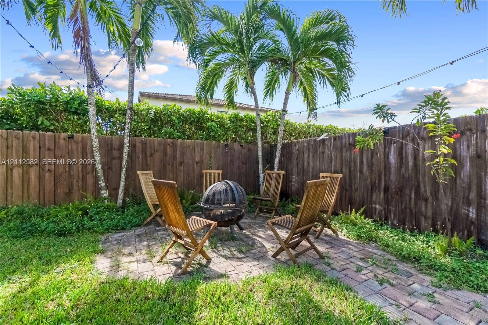 Backyard featuring a brick paver patio, wood privacy fencing, mature palm trees, and overhead string lighting