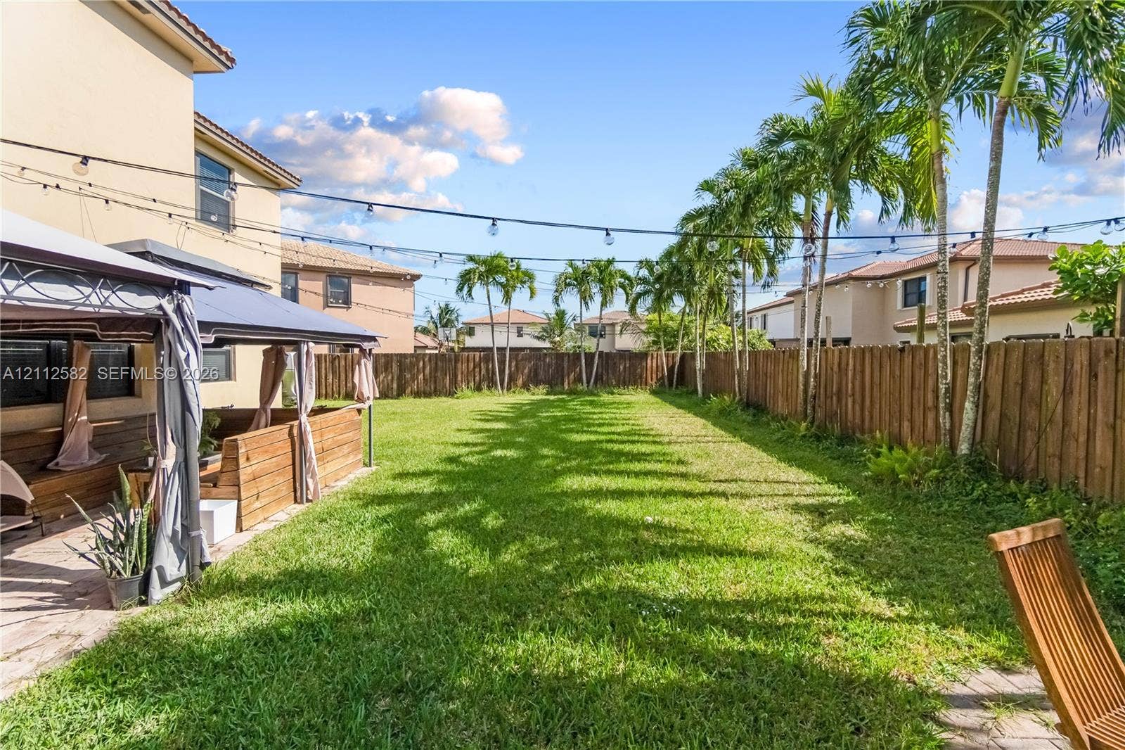 Expansive backyard featuring a manicured lawn, wood-panel fencing, mature palm trees, and a permanent metal-framed gazebo structure on a paved patio