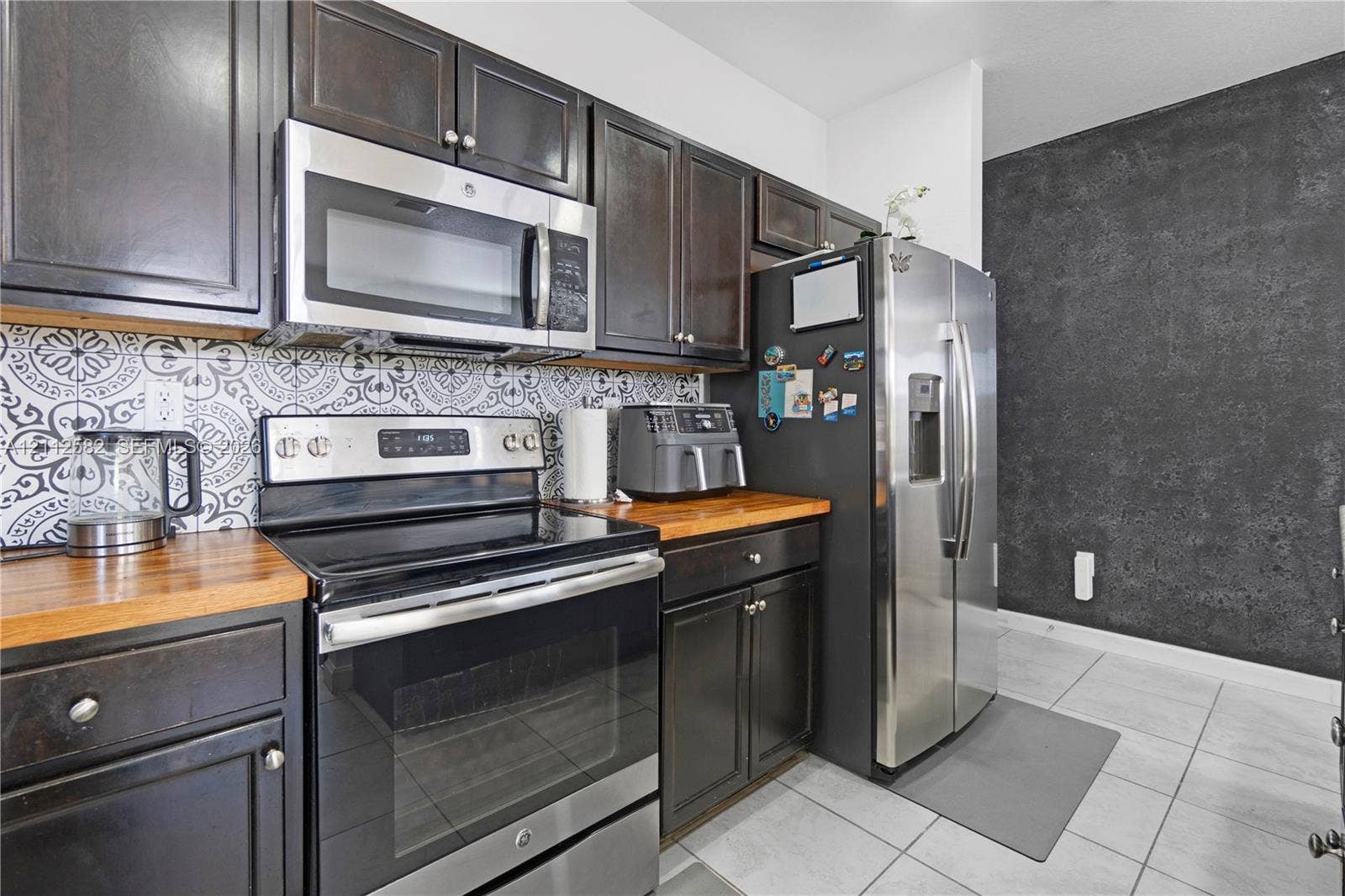 Kitchen featuring dark-finish cabinetry, butcher-block countertops, stainless steel appliances, decorative patterned backsplash, and tile flooring