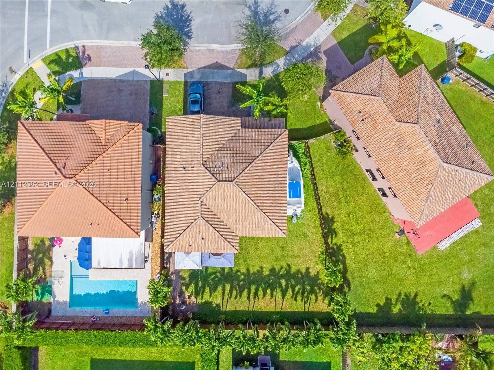 Aerial perspective featuring multi-pitched tile roofs, a private swimming pool with concrete decking, and mature palm landscaping within a fenced perimeter