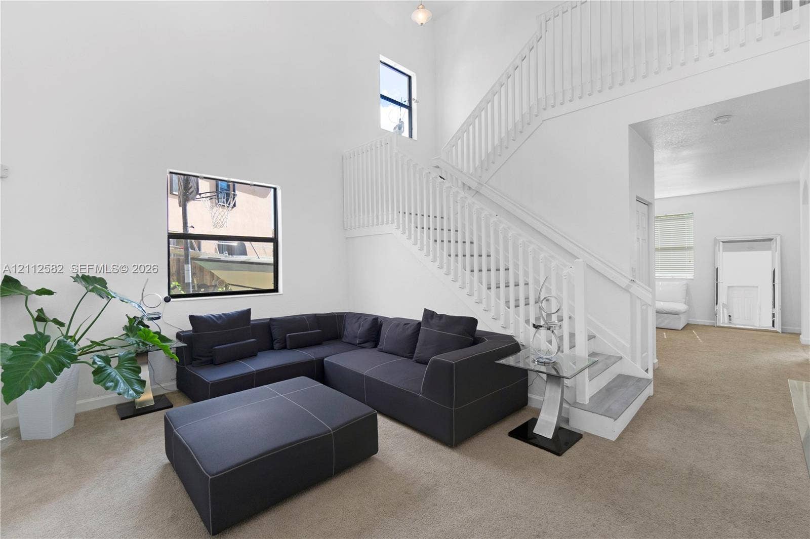 High-ceiling living area featuring a central staircase with white wooden balusters, large windows, and carpeted flooring