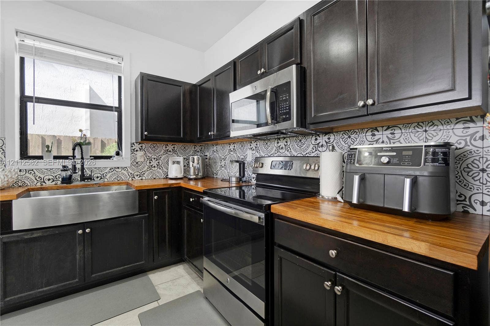 Kitchen featuring dark cabinetry, butcher block countertops, stainless steel farmhouse sink with a gooseneck faucet, stainless steel appliances, and patterned tile backsplash