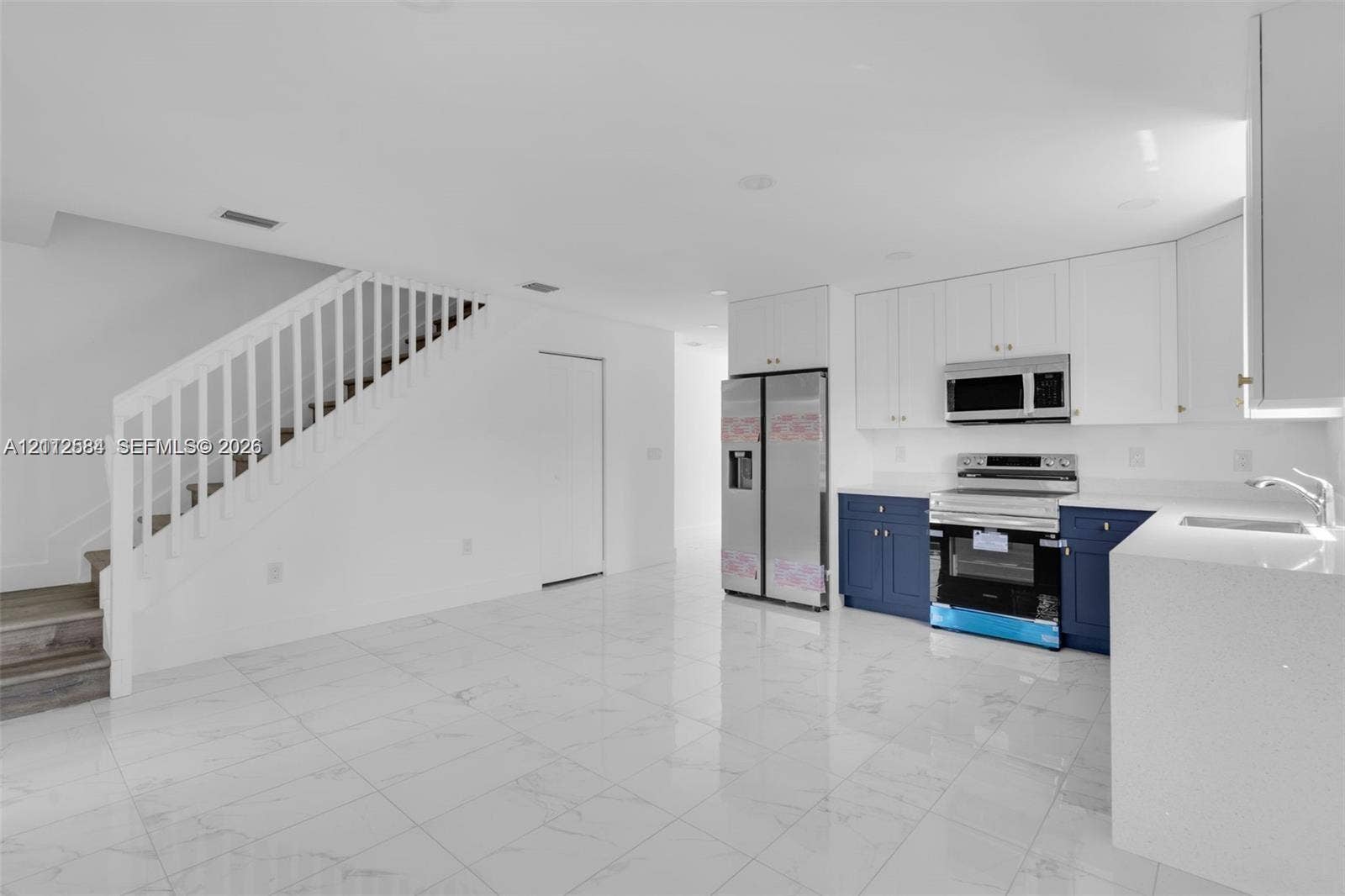 1629 NW 40th Street, Miami, FL 33142 Kitchen featuring white upper cabinetry, navy blue lower cabinetry, stainless steel appliances, and quartz countertops, complemented by white polished stone-finish flooring and a staircase with white wood railing