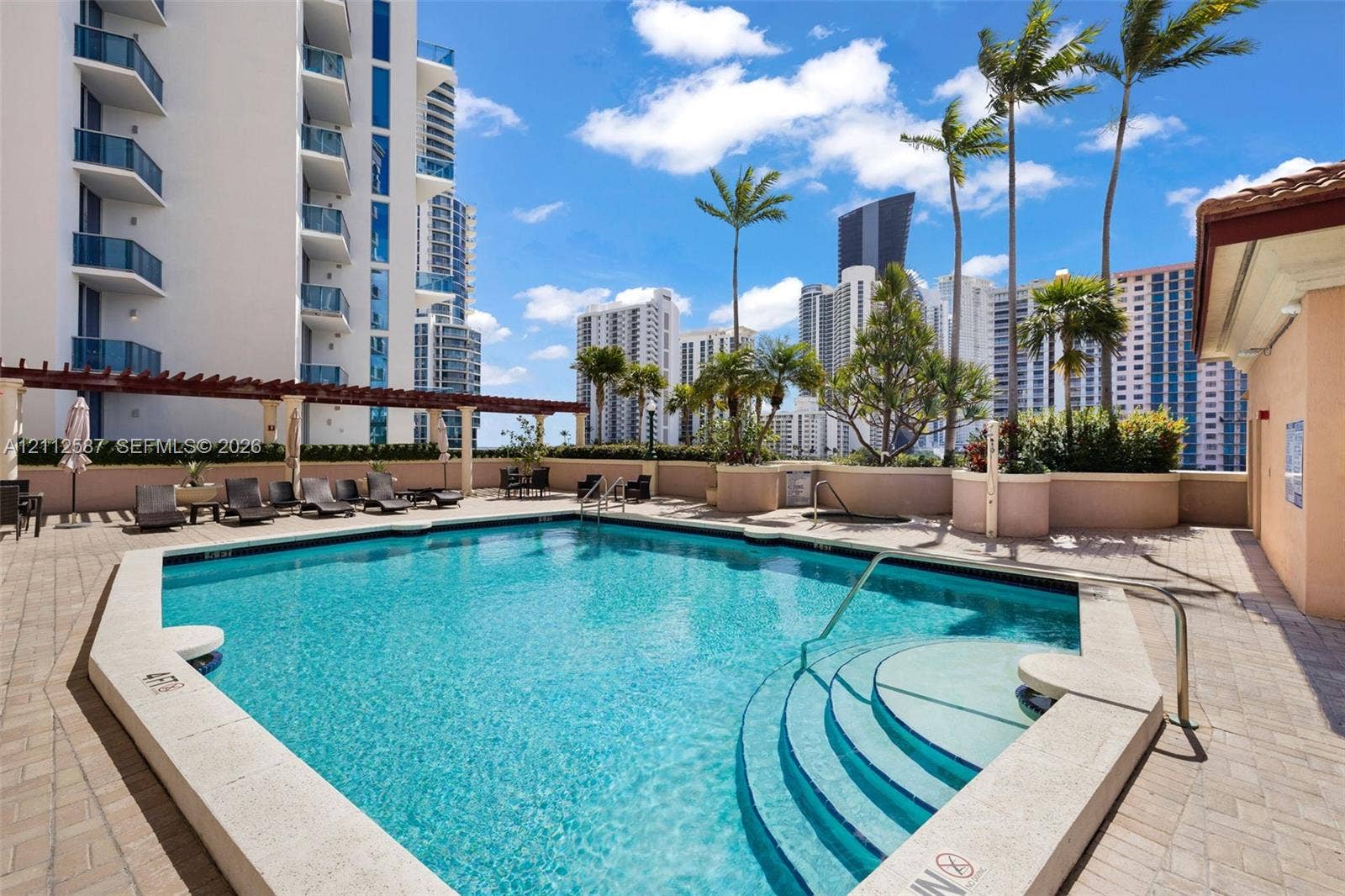 17555 NW Atlantic Boulevard, #1108, Sunny Isles Beach, FL 33160 Swimming pool with built-in stairs, perimeter stone coping, surrounding brick-paver terrace, wooden pergola structure, and mature palm landscaping