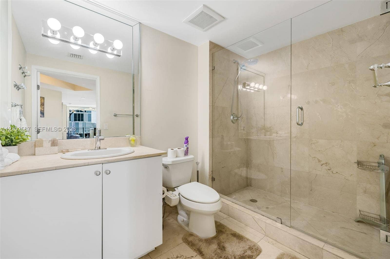 17555 NW Atlantic Boulevard, #1108, Sunny Isles Beach, FL 33160 Bathroom featuring a white vanity with an integrated sink, a full-width vanity mirror, a walk-in shower with a glass enclosure and stone-finish wall tile, and polished chrome hardware