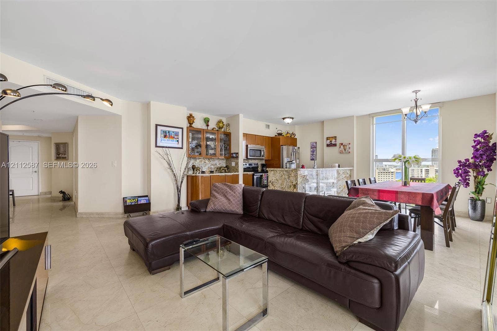 17555 NW Atlantic Boulevard, #1108, Sunny Isles Beach, FL 33160 Open-concept main living area featuring light-toned tile flooring, built-in wooden cabinetry with glass-front uppers, a kitchen island with stone countertop, and expansive floor-to-ceiling windows