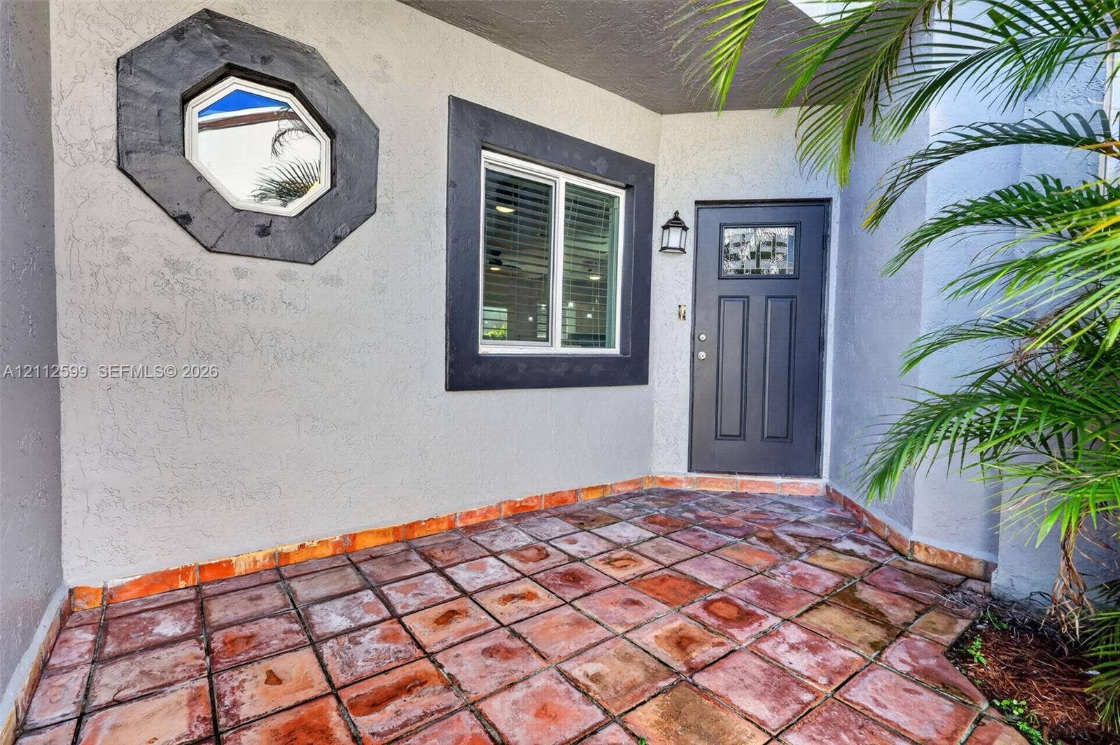 7974 NW 190th Terrace, Hialeah, FL 33015 Covered entry featuring terra-cotta tile flooring, a solid-core front door with decorative glass inlay, an octagonal accent window, and dark-finished exterior trim