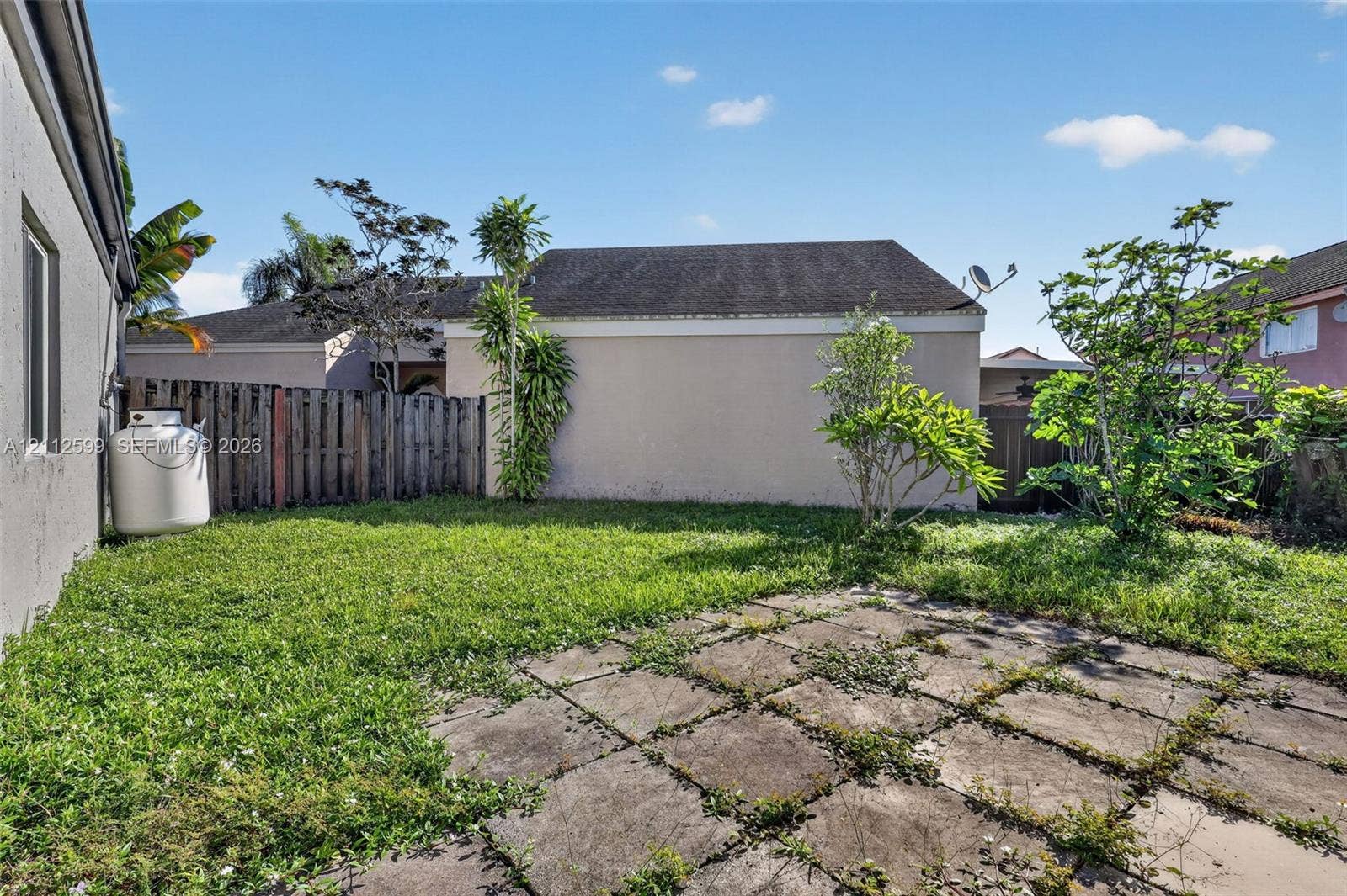 7974 NW 190th Terrace, Hialeah, FL 33015 Rear exterior grounds featuring a paved stone patio, lawn area, perimeter wood fencing, and mature landscaping