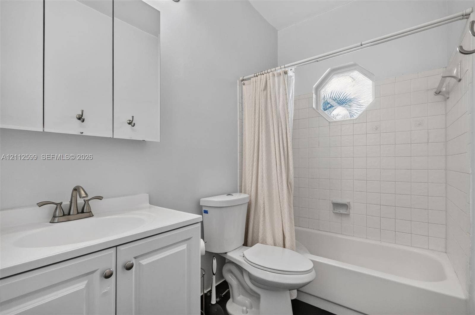 7974 NW 190th Terrace, Hialeah, FL 33015 Bathroom featuring a vanity cabinet with integrated countertop, octagonal window, white ceramic tile tub surround, and a wall-mounted mirrored medicine cabinet