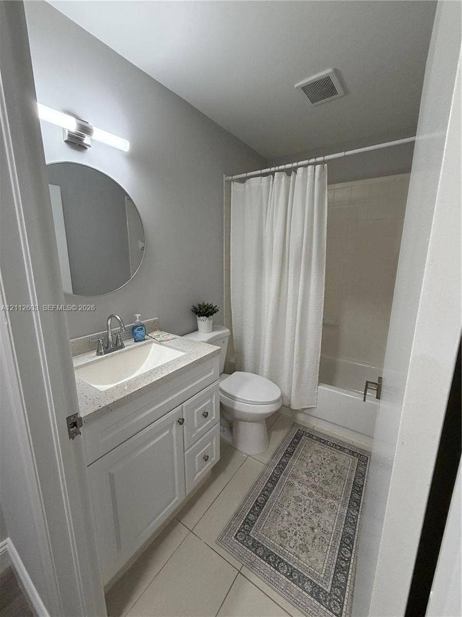 White cabinetry vanity with a speckled countertop, integrated basin, brushed nickel faucet, round wall-mounted mirror, and tile flooring
