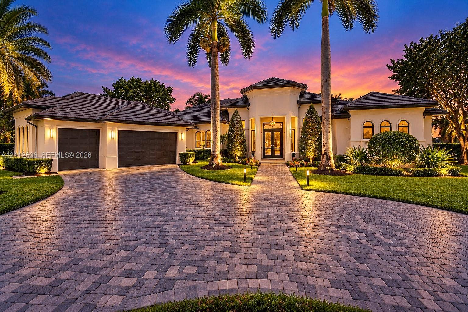 Stucco exterior featuring a multi-gabled tile roof, paver driveway, double-door entryway, and integrated landscape lighting