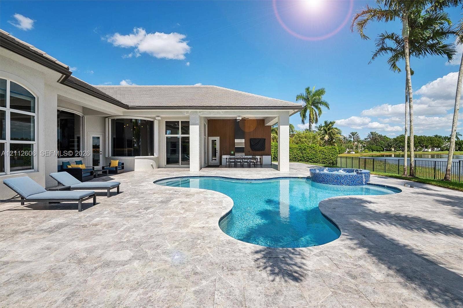 Outdoor living area featuring a paved stone pool deck, a free-form swimming pool with an integrated spa, a covered patio with an outdoor kitchen, and a water backdrop
