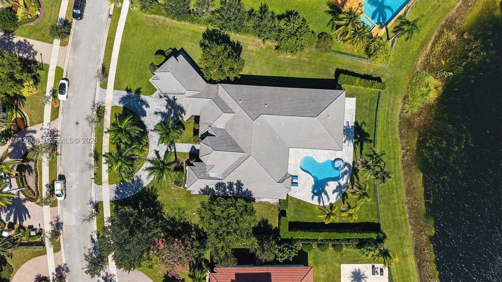 Aerial perspective of a property featuring a multi-pitched roofline, paved circular driveway, private swimming pool, and water frontage on a landscaped lot