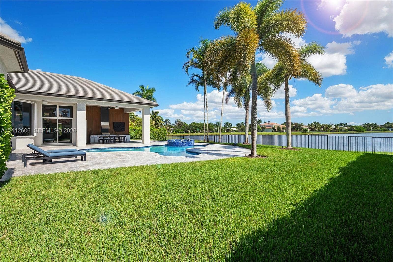 Rear exterior featuring a covered patio, paved pool deck, swimming pool with integrated spa, manicured lawn, and water frontage