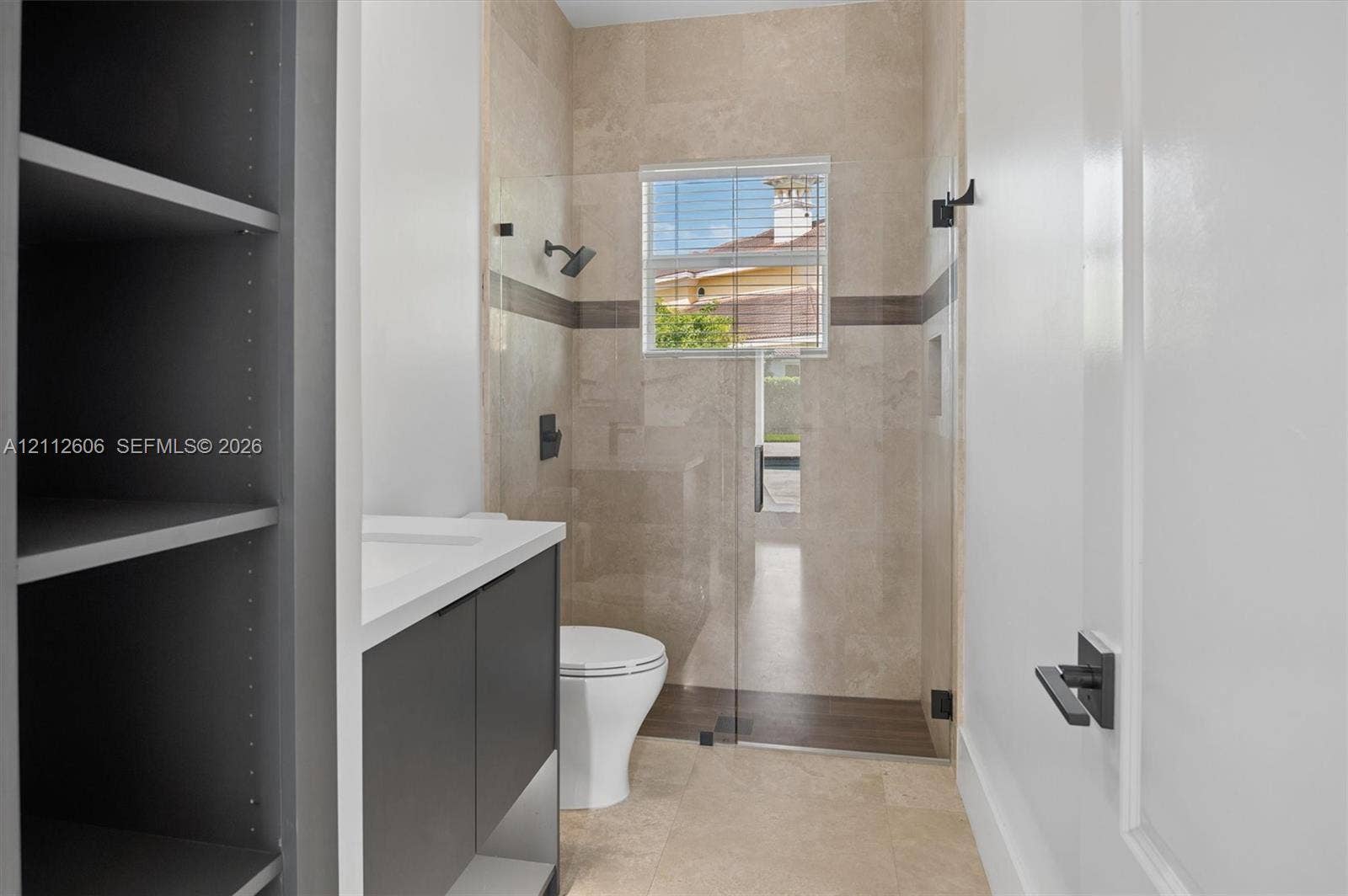 Bathroom featuring natural stone tile flooring, a glass-enclosed shower with contrasting horizontal tile inlay, a dark-finish vanity with a white countertop, and matte black hardware