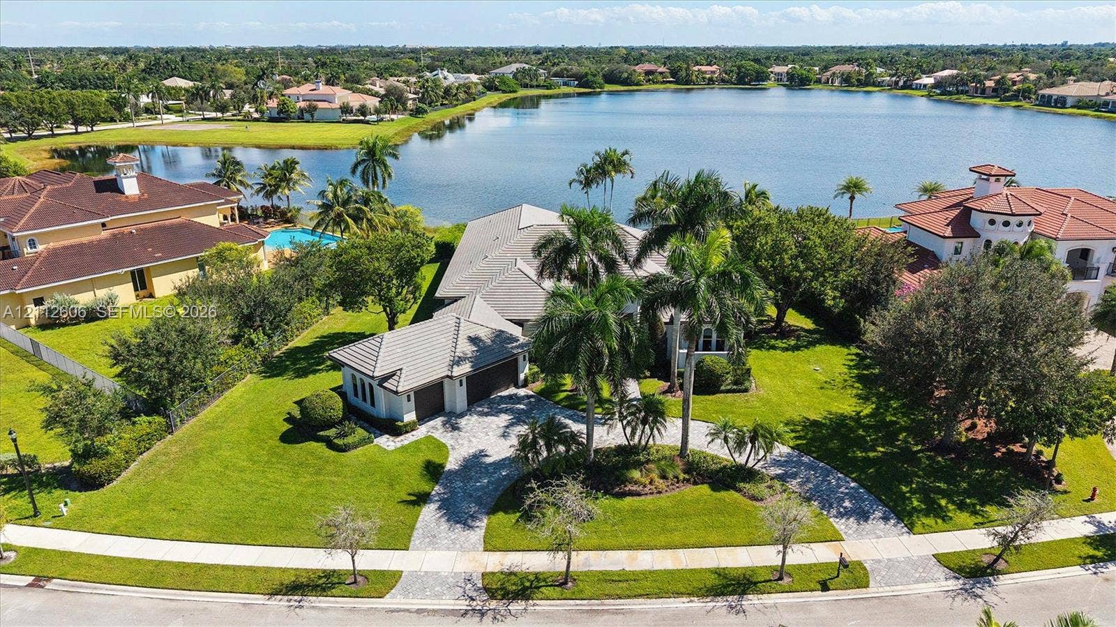 Aerial perspective featuring a multi-gabled residence with a concrete tile roof, a paved circular driveway, mature palm landscaping, and expansive water frontage