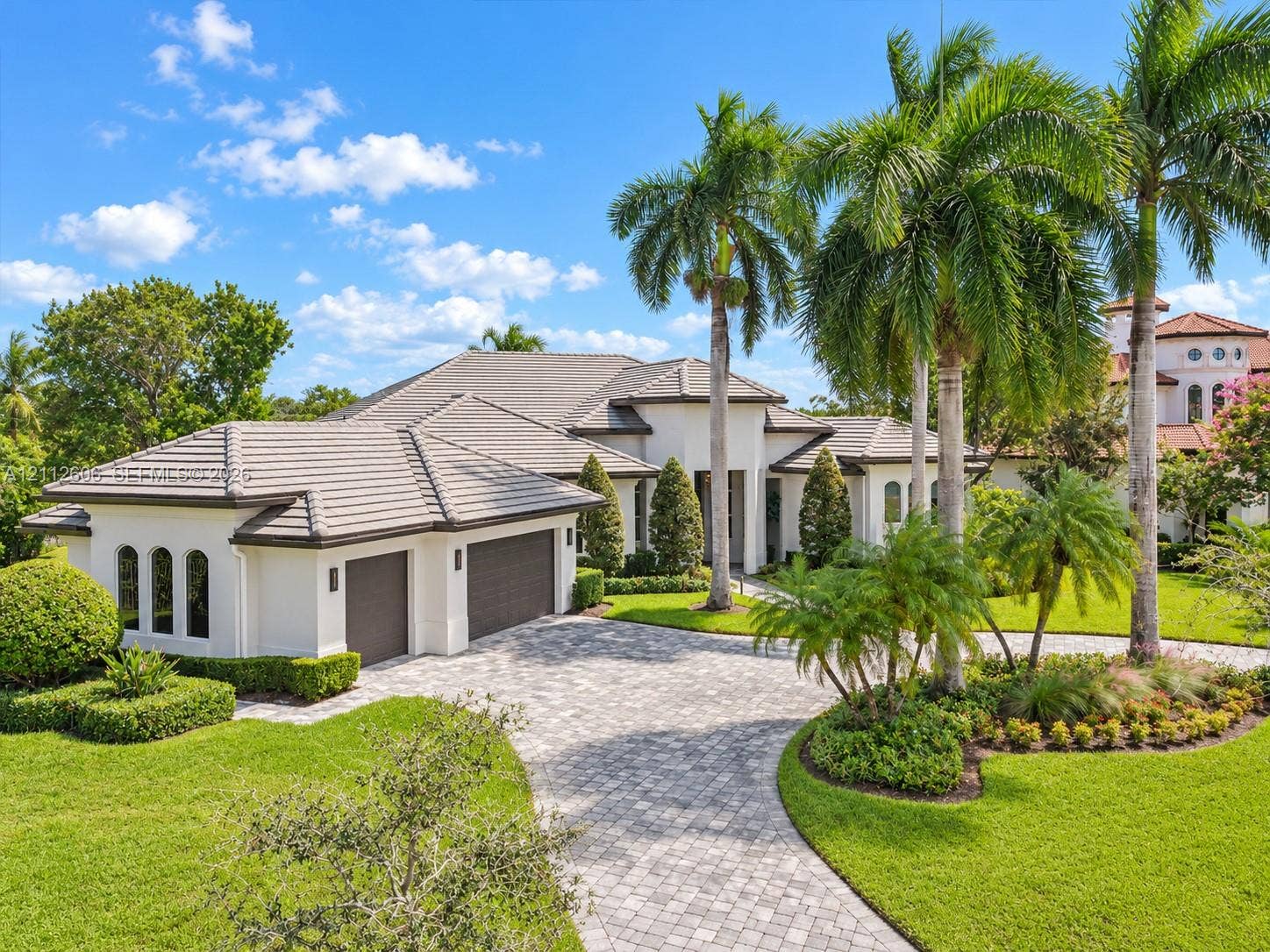 Stucco exterior with a multi-hipped tile roof, paver driveway, three-car garage, and mature palm landscaping