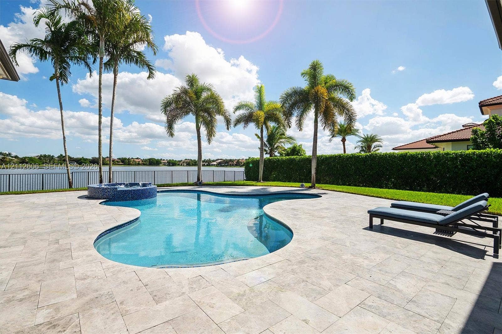 Freeform swimming pool with a raised spa, stone-paver decking, mature palm trees, and metal perimeter fencing overlooking a lake