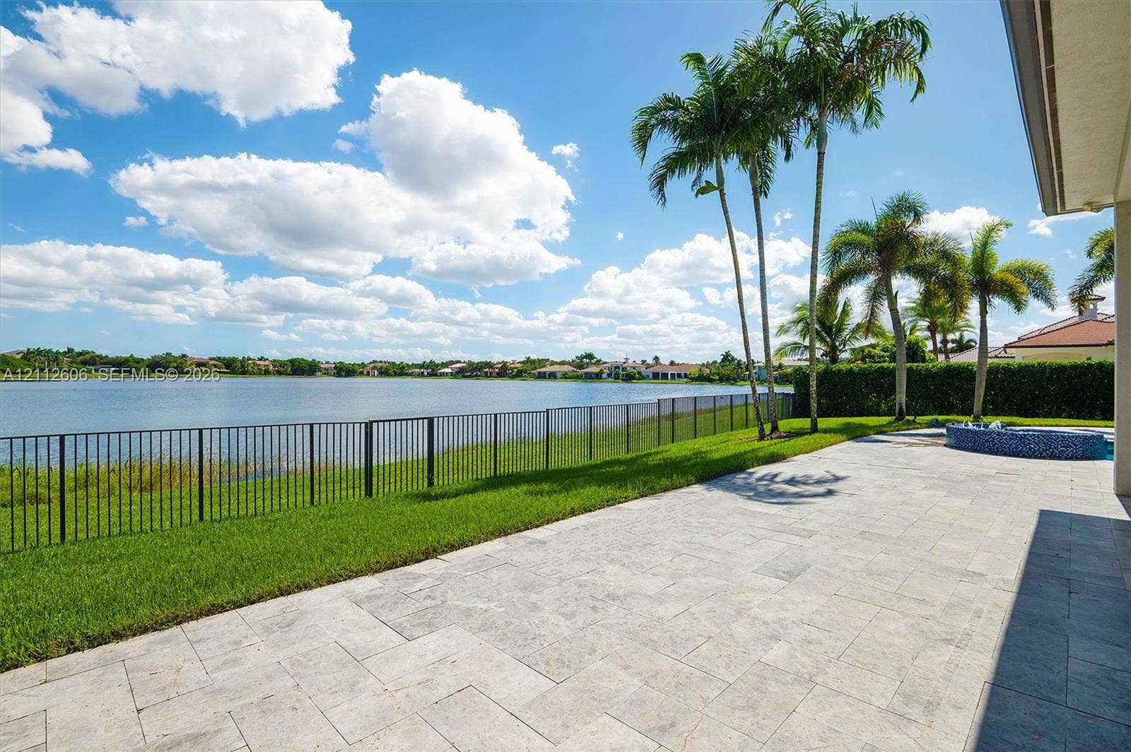 Paver patio featuring a tile-finished spa, metal perimeter fencing, manicured lawn, and mature palm trees with direct water frontage