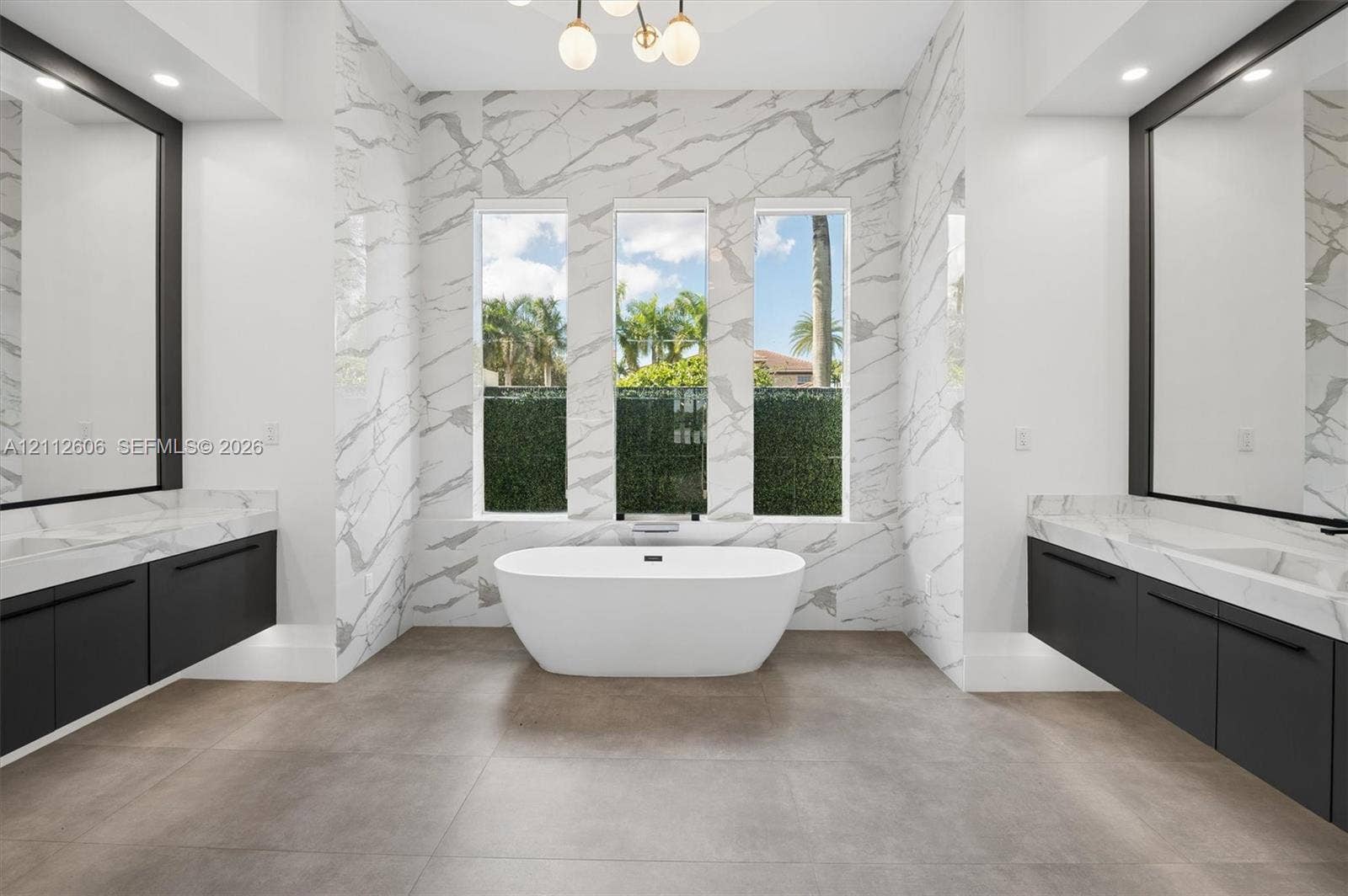 Freestanding soaking tub featuring large-format wall tiling, dual floating vanities with matte black hardware, framed wall-to-wall mirrors, and a modern multi-globe chandelier