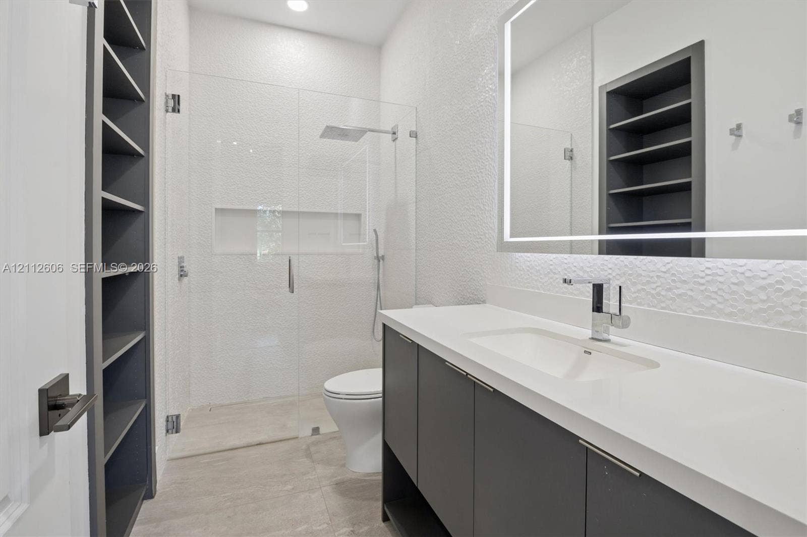 Floating vanity with solid-surface countertop and integrated sink, frameless glass-enclosed shower featuring built-in recessed niche, textured wall surfaces, illuminated vanity mirror, and integrated floor-to-ceiling shelving