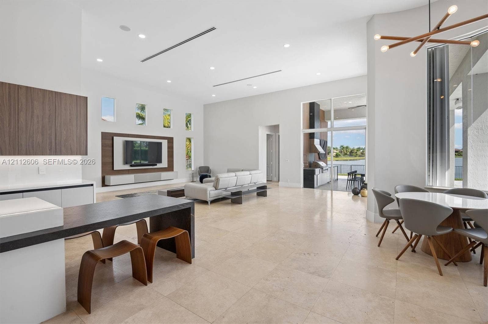 Open-concept interior featuring high ceilings, stone tile flooring, built-in media wall cabinetry, floor-to-ceiling glass paneling, and a contemporary multi-arm chandelier
