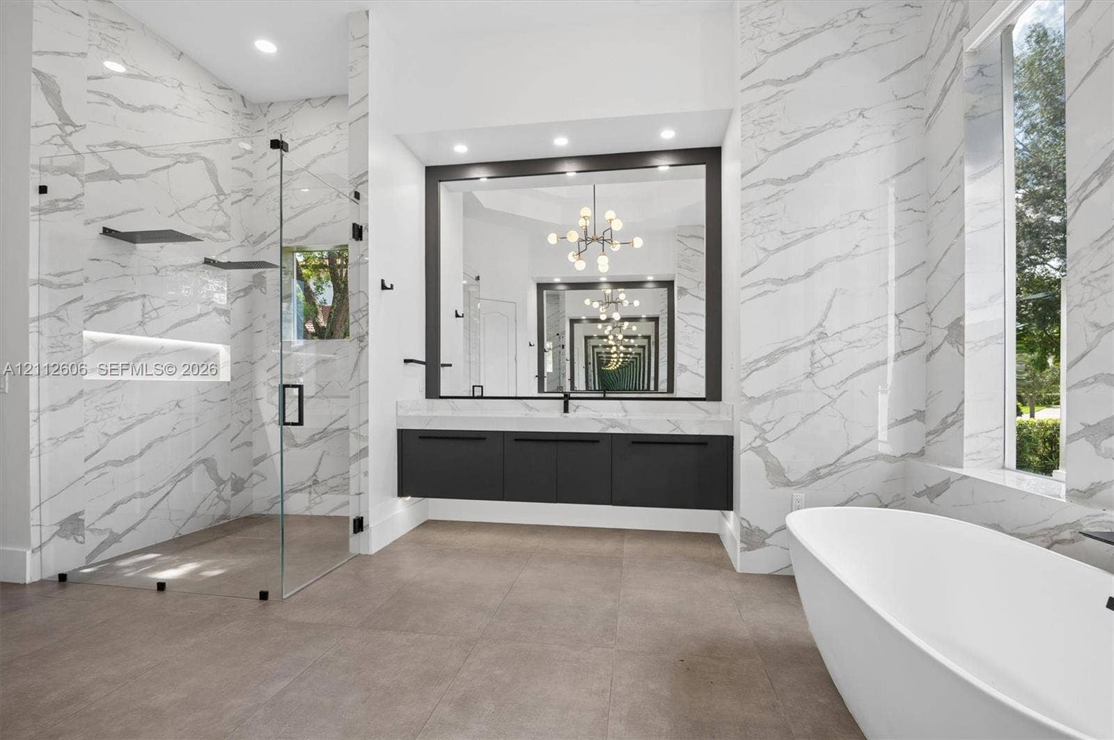 Walk-in glass shower with built-in shelving, floating vanity, large-format tile flooring, freestanding soaking tub, and stone-surround wall detailing