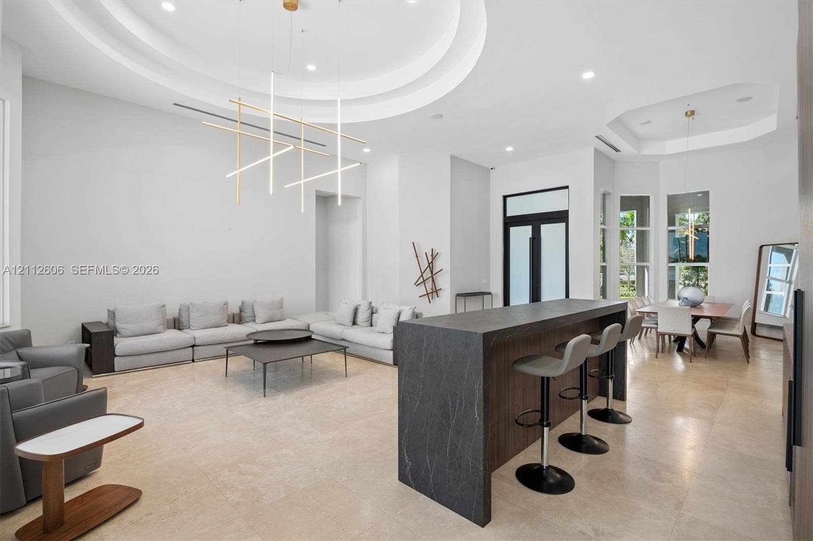 Open floor plan featuring high ceilings with recessed tray detailing, stone-finish flooring, a waterfall-edge kitchen island with dark cabinetry, and modern linear pendant lighting