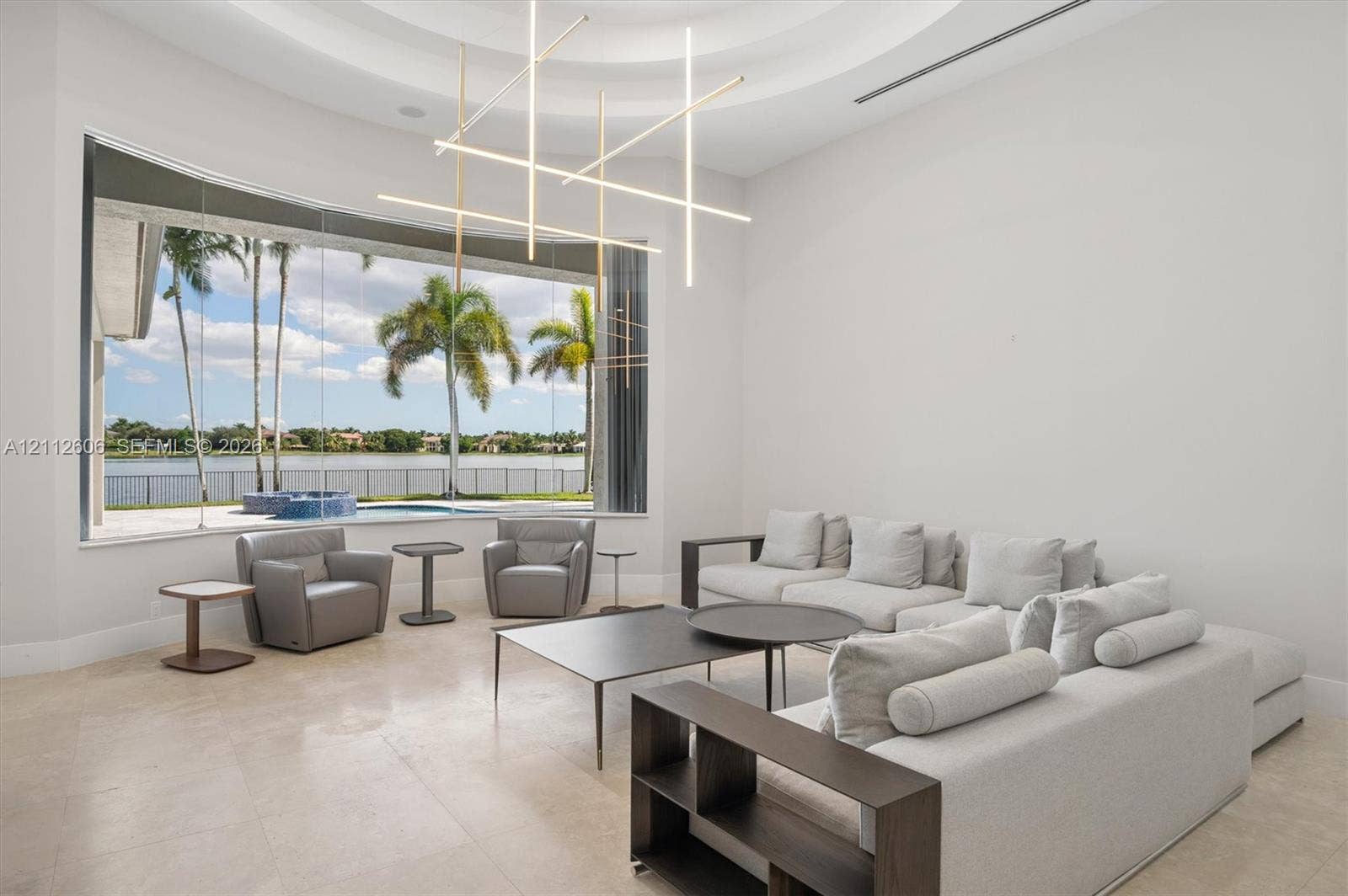 High-ceiling living area featuring large-format stone flooring, a circular tray ceiling, modern linear geometric chandelier, and floor-to-ceiling windows with waterfront scenery