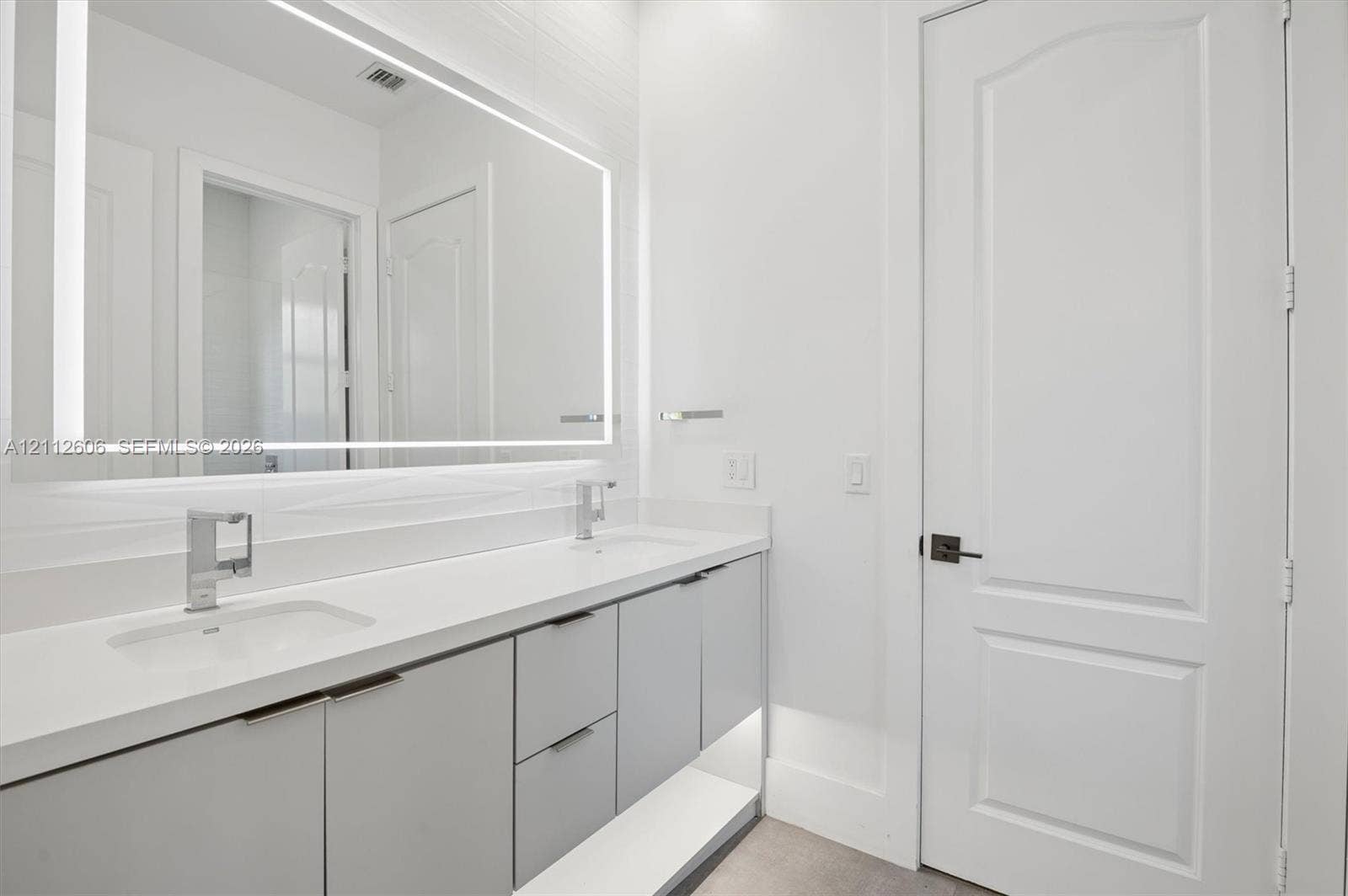 Double vanity featuring a light-toned countertop with integrated dual sinks, sleek cabinetry with minimalist hardware, and a large LED-backlit mirror