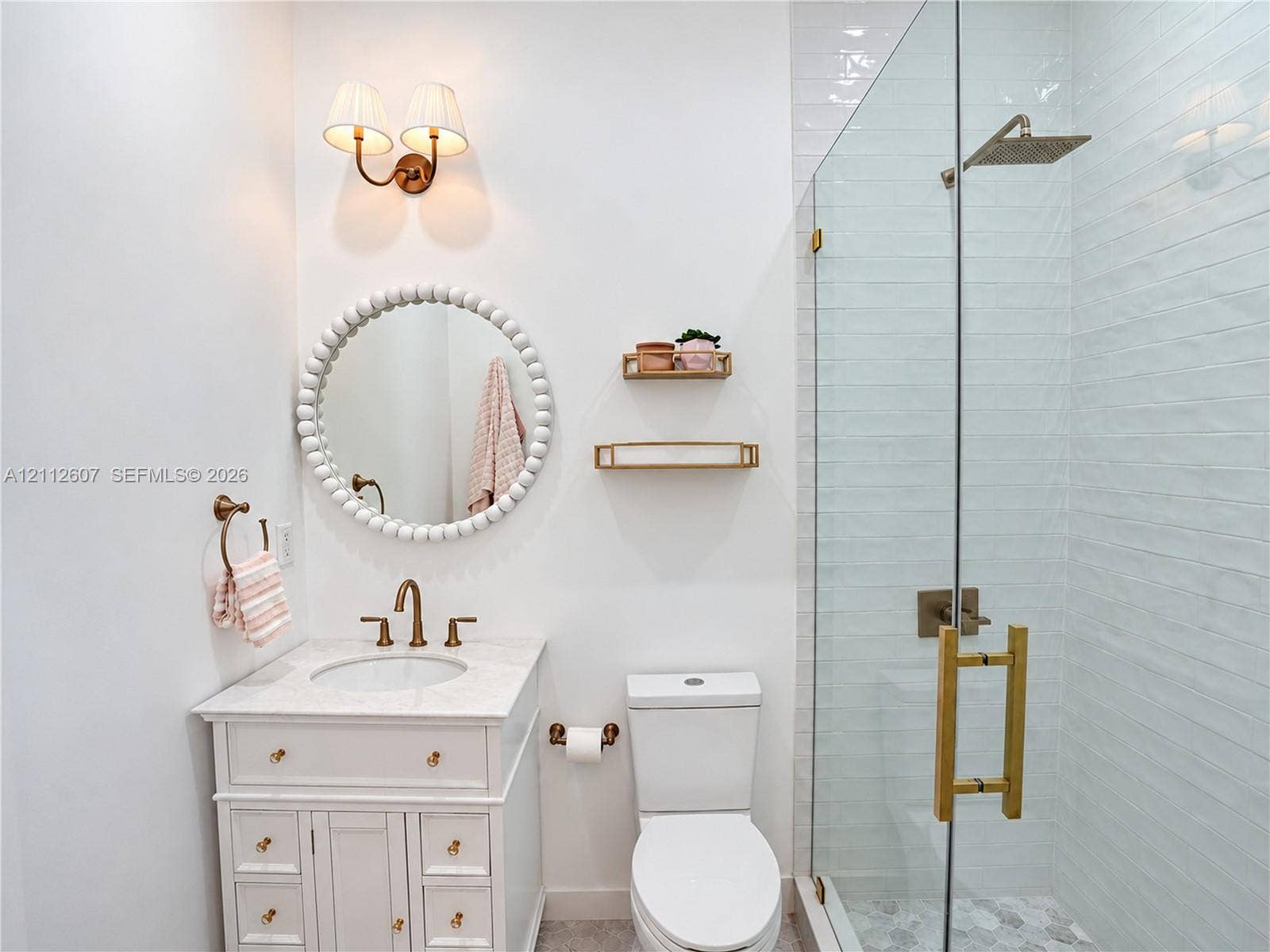 14443 S Jockey Circle S, Davie, FL 33330 White vanity cabinet with a stone countertop, wall-mounted dual-bulb light fixture, circular bead-detail mirror, and a glass-enclosed shower with a rainfall showerhead and polished metal hardware
